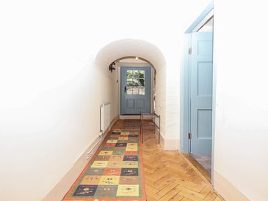 A hallway with a patterned runner rug wooden parquet floor white walls light blue door at the end and a small metal rack near the door at Aluric's Hall in Porlock