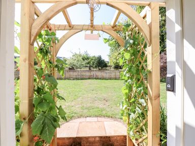 A wooden garden arch with climbing plants leading to a grassy yard with a wooden fence and trees at Aluric's Hall in Porlock