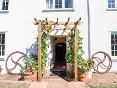 An entrance with a wooden pergola covered in plants and potted flowers by a white building at Aluric's Hall in Porlock