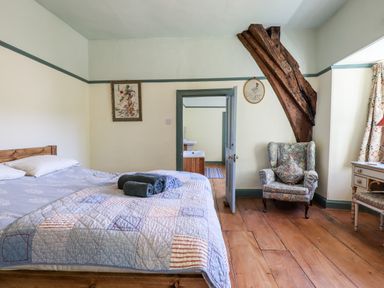 A bedroom with a bed towels on it an armchair wooden beam hardwood floor and doorway to a bathroom at Aluric's Hall in Porlock