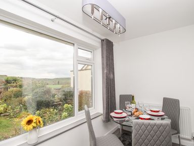 A dining area with a glass table set with plates bowls and wine glasses beside a window showing outdoor greenery at Rosamond in Shipton Gorge