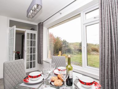 A dining area with a glass table set with plates bowls glasses bread and a bottle by a large window with grey curtains at Rosamond in Shipton Gorge