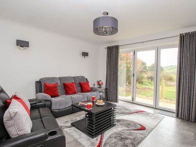 A living room with sofas and red cushions next to large glass doors with a view of greenery at Rosamond in Shipton Gorge