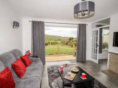A living room with a gray sofa and red cushions a black coffee table with mugs and treats and a view of green fields through glass doors at Rosamond in Shipton Gorge