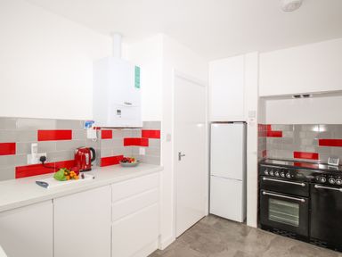A kitchen with white cabinets, a black stove, white refrigerator, red and gray tiled backsplash, and a countertop with a cutting board and kettle at Rosamond in Shipton Gorge