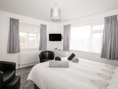 A bedroom with a double bed with rolled towels two black armchairs a wall mounted TV and gray curtains at Rosamond in Shipton Gorge
