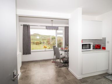 A dining area with two chairs and a table near a window with curtains and a kitchen counter with a microwave and toaster at Rosamond in Shipton Gorge