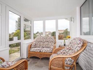 A sunroom with wicker chairs and loveseat with printed cushions a small table with wine glasses and sunflowers at Rosamond in Shipton Gorge