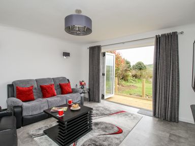 A living room with a gray sofa with red pillows a black coffee table with red mugs and pastries gray curtains open to a garden at Rosamond in Shipton Gorge