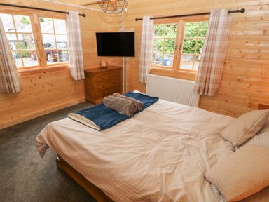 A bedroom with a bed covered with blankets and towels two windows with curtains a wooden chest of drawers and a wall-mounted television at Bryn Derwen Lodge in Bethesda