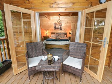 A wooden bedroom with a bed and bedside tables viewed through open glass doors onto a deck with two wicker chairs and a small table with coffee items at Bryn Derwen Lodge in Bethesda