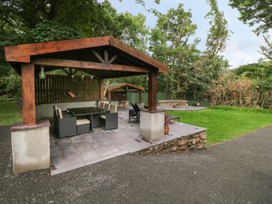 An outdoor covered seating area with table and chairs near a grassy area and fire pit at Bryn Derwen Lodge in Bethesda
