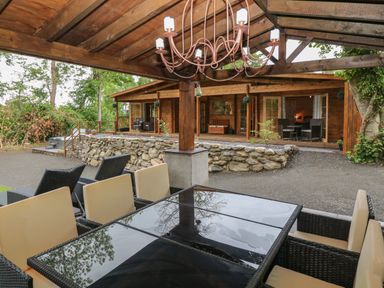 An outdoor seating area with a glass table and cushioned chairs under a wooden pergola next to a stone wall and wooden cabin at Bryn Derwen Lodge in Bethesda