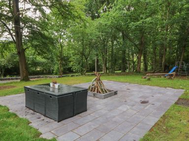 An outdoor patio with a dining table and chairs a fire pit with stacked logs and a playground slide in a grassy area surrounded by trees at Bryn Derwen Lodge in Bethesda
