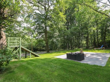 A grassy outdoor area with a wooden zipline platform and a paved seating area with firewood and chairs surrounded by trees at Bryn Derwen Lodge in Bethesda