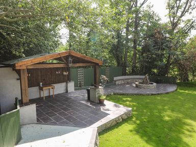 An outdoor patio with a wooden covered bar area and a circular stone fire pit with curved bench seating at Bryn Derwen Lodge in Bethesda