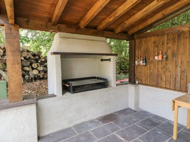 An outdoor covered barbecue area with a built-in grill and bottles on shelves at Bryn Derwen Lodge in Bethesda