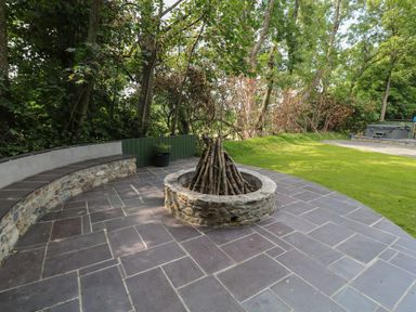 An outdoor patio area with a stone fire pit with stacked logs and curved stone seating surrounded by trees at Bryn Derwen Lodge Bethesda
