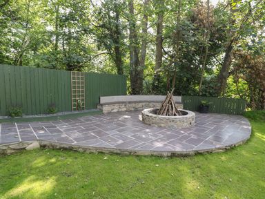 An outdoor patio with stone fire pit surrounded by wooden benches and green fence at Bryn Derwen Lodge in Bethesda