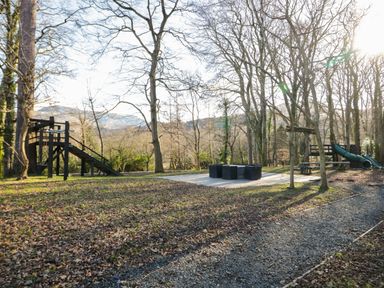 A children's play area with swings slides and a seating area in a wooded park at Bryn Derwen Lodge in Bethesda