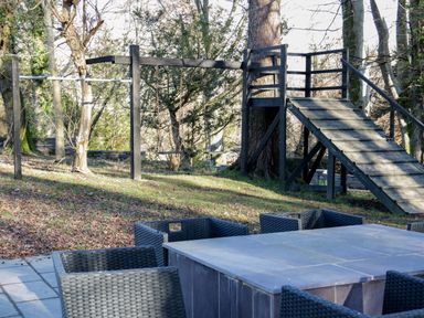 An outdoor patio area with a gray table and six woven chairs next to a wooden playground structure with swings at Bryn Derwen Lodge in Bethesda