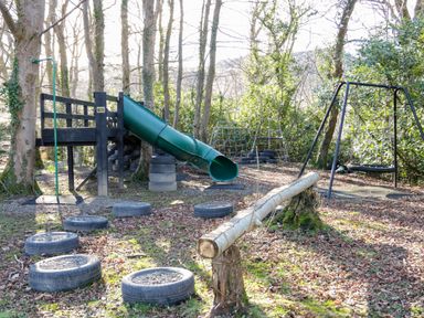 An outdoor playground with a wooden climbing structure and slide tire stepping stones a swing and climbing frames in a wooded area at Bryn Derwen Lodge in Bethesda