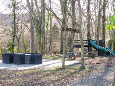 An outdoor area with a seating set and a playground with a slide and swing among trees at Bryn Derwen Lodge in Bethesda