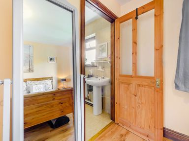 A view of a wooden bed and nightstand in a bedroom reflected in a mirror and a doorway showing a bathroom with a pedestal sink at Covert Farm Cwtch in Jeffreyston