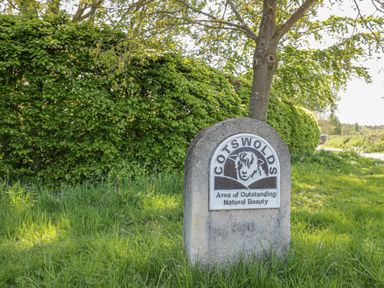 A stone marker with a sign reading Cotswolds Area of Outstanding Natural Beauty standing on grass near a tree and hedge at Spring Cottage in Alkerton near Banbury