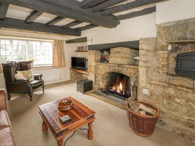 A living room with a stone fireplace burning fire a wooden table leather armchair and a television at Spring Cottage in Alkerton near Banbury