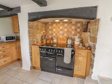 A kitchen with a black stove set in a stone alcove with wooden cabinets and tiled backsplash at Spring Cottage in Alkerton near Banbury