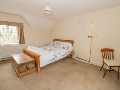 A bedroom with a wooden bed with white and floral bedding a wooden chest at the foot of the bed a wooden chair with a cushion a window with curtains and a radiator under the window and a closed door at Spring Cottage in Alkerton near Banbury