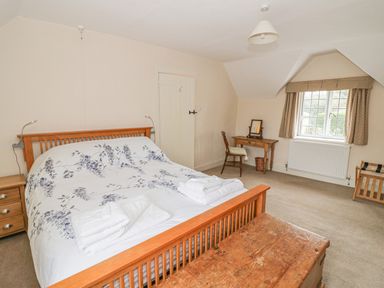 A bedroom with a wooden bed and white floral bedding a wooden chest at the foot of the bed a wooden desk and chair near a window with beige curtains at Spring Cottage Alkerton near Banbury