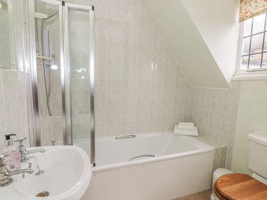 A bathroom with a bathtub with glass shower doors a white sink with soap next to a toilet with a wooden seat and a window at Spring Cottage in Alkerton near Banbury