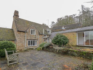 An outdoor courtyard with stone paving a wooden bench and a stone building with windows and foliage at Spring Cottage Alkerton near Banbury