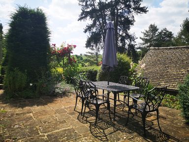 An outdoor patio with a metal table and six chairs with a closed umbrella surrounded by trees and bushes at Spring Cottage in Alkerton near Banbury