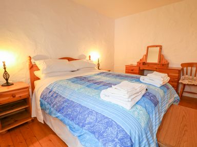 A bedroom with a double bed with blue bedding wooden nightstands lamps dressing table and a chair at Quiet Waters Cottage in Loughan near Coleraine
