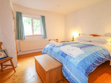 A bedroom with a double bed covered by a blue patterned quilt and folded towels on top a wooden chest at the foot of the bed a wooden chair with a cushion by the window with curtains at Quiet Waters Cottage in Loughan near Coleraine