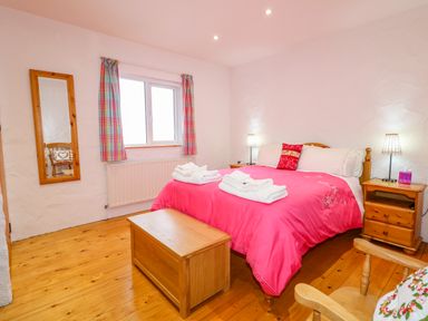 A bedroom with a wooden bed covered with a pink bedspread two wooden nightstands with lamps a wooden chest at the foot of the bed a mirror on the wall and a window with checkered curtains at Quiet Waters Cottage in Loughan near Coleraine