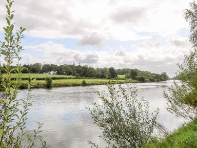 A river with green bushes and trees along the banks and a white house in the distance at Quiet Waters Cottage in Loughan near Coleraine