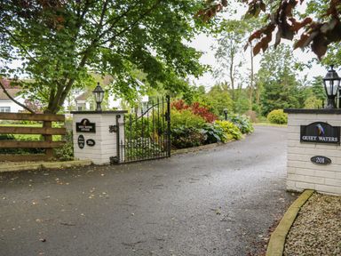 A driveway entrance with a black iron gate and white brick pillars with signs reading Quiet Waters Cottage at Quiet Waters Cottage in Loughan near Coleraine