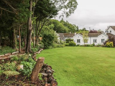 A grassy lawn with trees and plants near white houses at Quiet Waters Cottage in Loughan near Coleraine