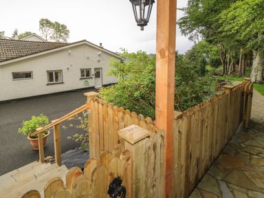 A wooden fence and stairs leading to a paved walkway with a white building and green trees in the background at Quiet Waters Cottage in Loughan near Coleraine