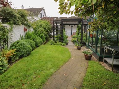 A garden with a paved walkway leading to a wooden pergola and a greenhouse on the right at Quiet Waters Cottage in Loughan near Coleraine