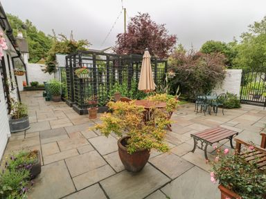 An outdoor patio area with stone paving wooden tables and chairs potted plants a black trellis structure and a metal gate at Quiet Waters Cottage in Loughan near Coleraine