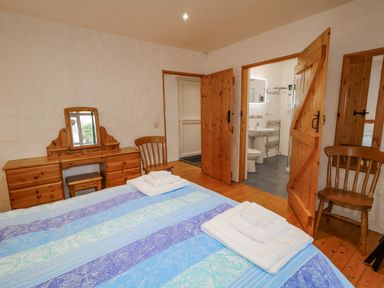 A bedroom with a bed covered by a blue patterned blanket two wooden chairs a wooden dressing table with a mirror wooden doors leading to an en suite bathroom at Quiet Waters Cottage in Loughan near Coleraine