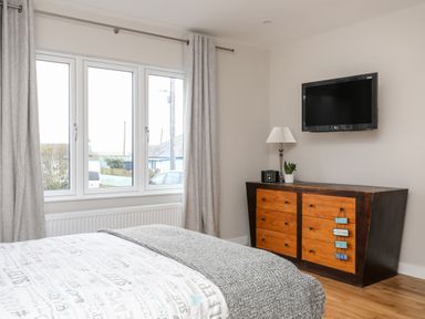 A bedroom with a bed next to a window and a wooden dresser with a lamp and a television on the wall at Laneton in Rhosneigr