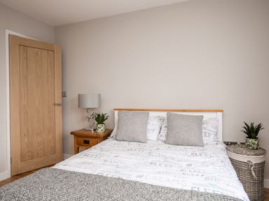 A bedroom with a bed with gray pillows and patterned bedding two plants on wooden and wicker bedside tables at Laneton in Rhosneigr