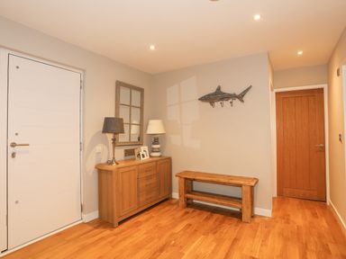An entryway with wooden floor a white door a wooden cabinet with two lamps and photo frames a wooden bench and a wooden fish wall hook at Laneton in Rhosneigr