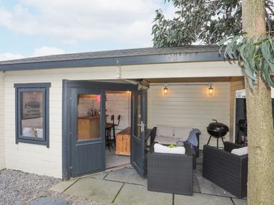 An outdoor patio area with black wicker seating and beige cushions next to an open door leading to a room with bar stools at Laneton in Rhosneigr